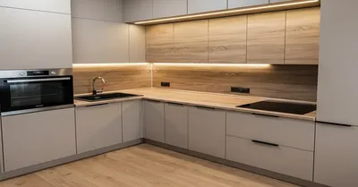 Интерьер современной кухни с матовыми бежевыми шкафами, фартуком из МДФ дерево и столешницей. Угловая планировка, встроенная духовка и светодиодная подсветка рабочей зоны под верхними шкафами.