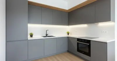 Вид на современную Г-образную кухню с серыми матовыми фасадами, белой бесшовной столешницей и белым кухонным фартуком. Под верхними шкафами установлена ровная линейная подсветка, встроен черный духовой шкаф и мойка с черным смесителем.